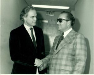 Jay J Armes in a plaid suit shakes hands with Marlon Brando in a dark suit. Main Course.